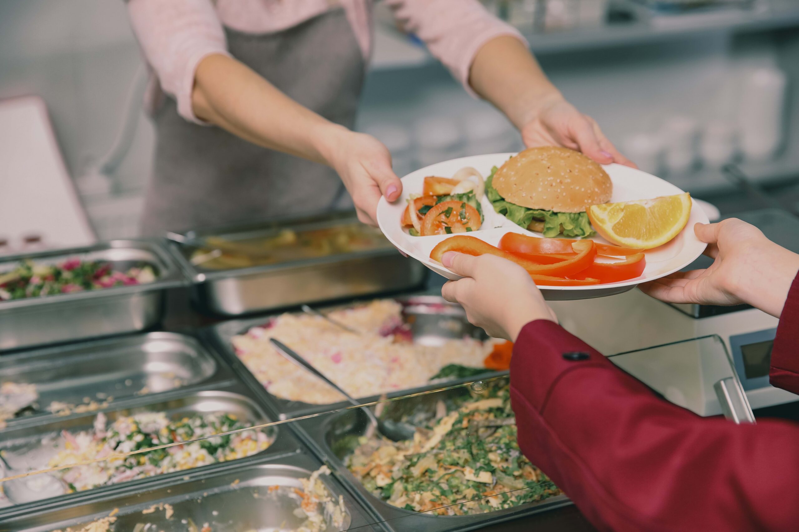 A pair of hands receives a round lunch plate with a sandwich, orange slices, pepper slices, and salad from another pair of hands. The transfer of the plate is happening above a food bar with some casseroles in serving trays.