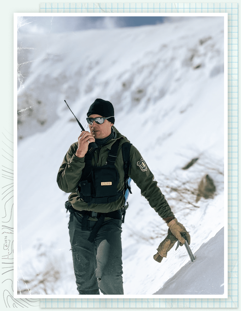 A clean shaven man wearing an olive green U.S. Forest Service jacket, gray-green snow pants, thick work gloves, a black beanie, sunglasses, and two equipment packs across his chest leans on a metal stake embedded in a snowy embankment with one hand while raising a walkie talkie to his mouth with the other hand.