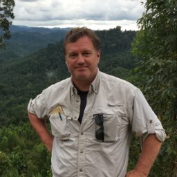 Scott Paul poses in a white button-up shirt in front of a jungle background. Scott Paul poses in a white button-up shirt in front of a jungle background.