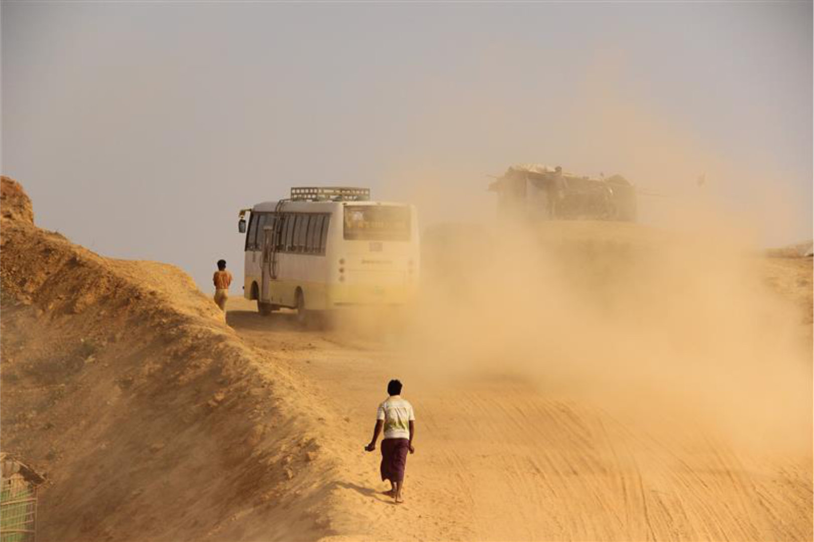Rohingya Camp, Cox's Bazar. Dirt road with dust flying up behind vehicles onto boy walking up the hill.
