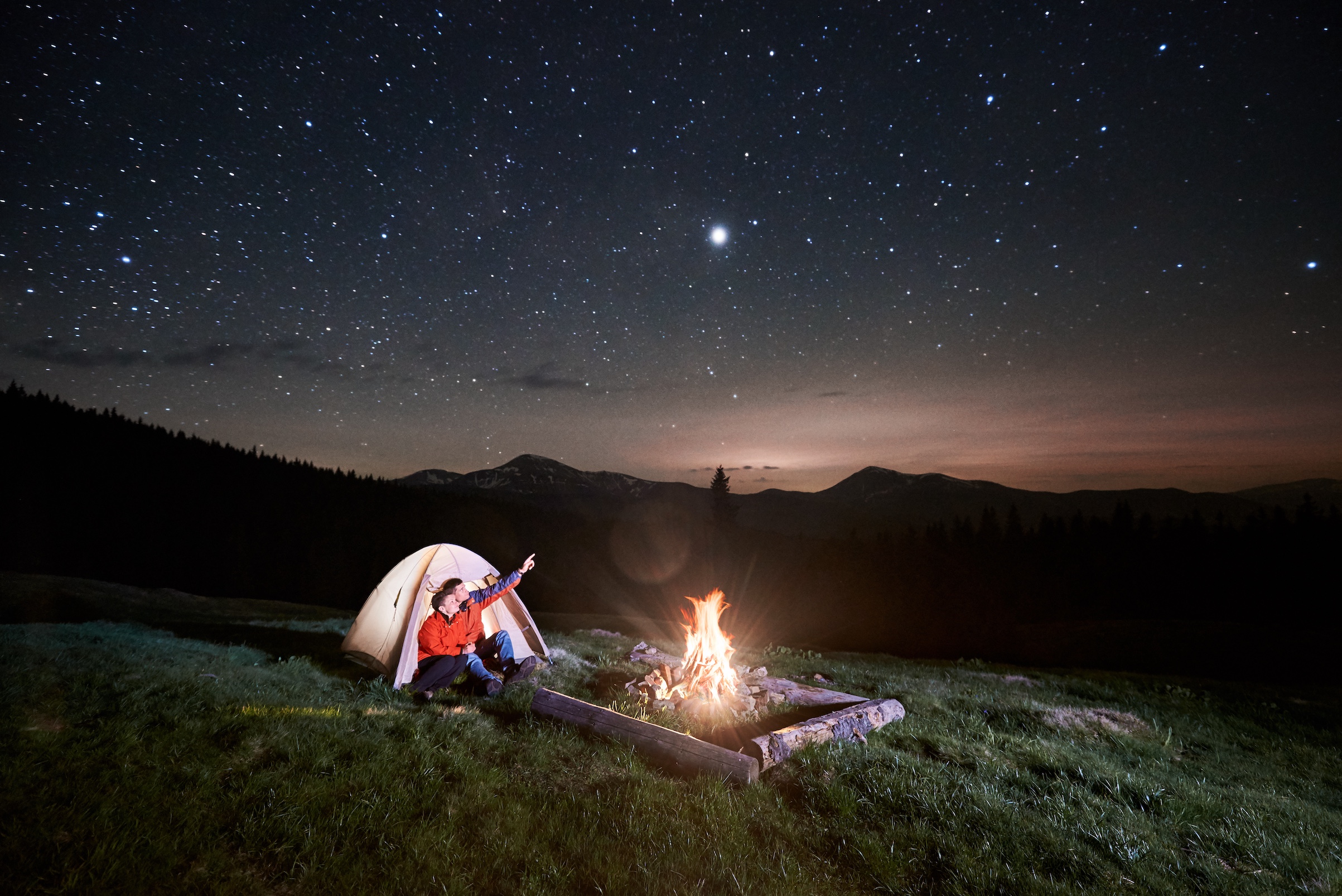Two people sitting outside a tent in front of a campfire, one of them is pointing up at the night sky filled with stars.