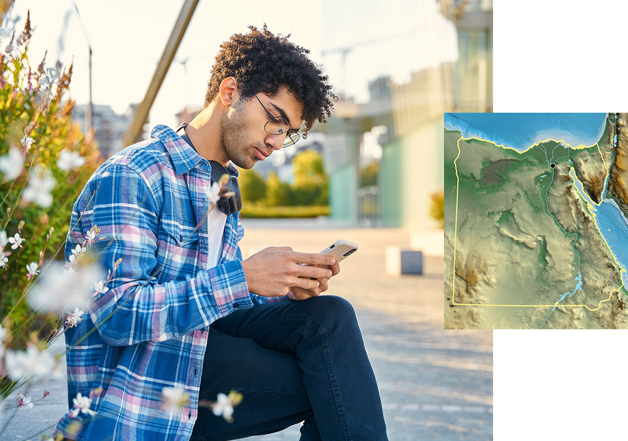 A college student sitting in a landscaped campus quad looking at a cell phone screen, overlaid with a small terrain map
