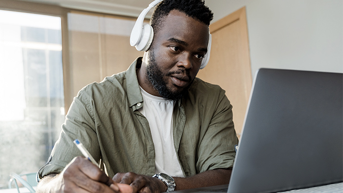 University student wearing headphones looking at a laptop while writing notes