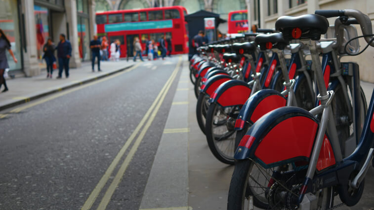 A bicycle share next to a street, with pedestrians and double-decker buses in the distance 