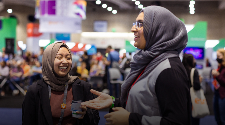 Two laughing conference attendees in headscarves standing in a busy expo hall 