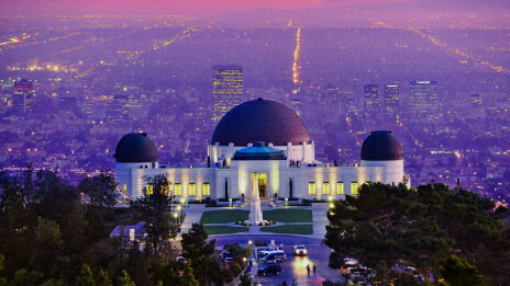 A large white observatory with several domes lit up for sunset with a sprawling purple-hued city in the background