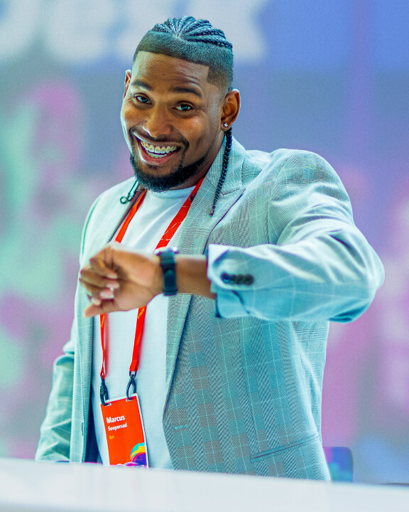 A person in a light blue checked blazer and red conference lanyard smiling up at the camera in the middle of checking their watch