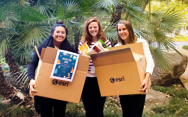 Three smiling people in front of a small palm tree holding large cardboard boxes printed with Esri’s logo