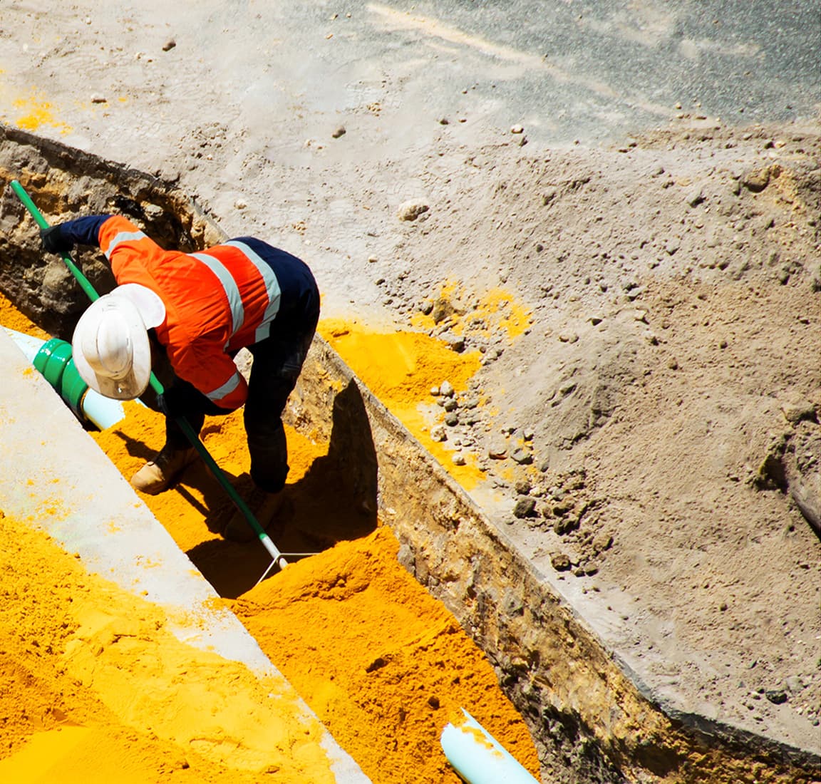 A utility worker in an orange vest and white sunshade hard hat shoveling yellow dirt in a trench