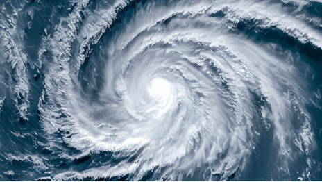 A satellite image of a hurricane with white clouds spiraling around a dense eye against a blue ocean background.