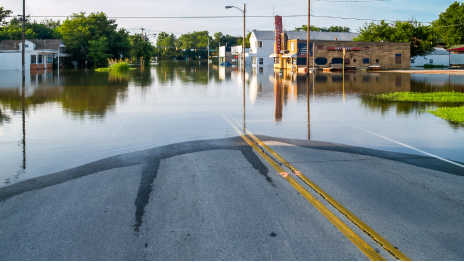 A street view photograph shows a flooded residential neighborhood with water covering the road and nearby lawns.