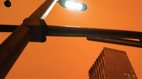 On an orange background, an up-close image of a black light pole with a white light hangs over a tall building