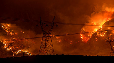 Blazing red and orange wildfires with a large black telephone pole and powerlines in the foreground