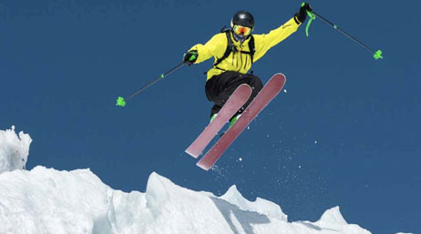 A snow skier in a yellow jacket is mid-jump off a large embankment