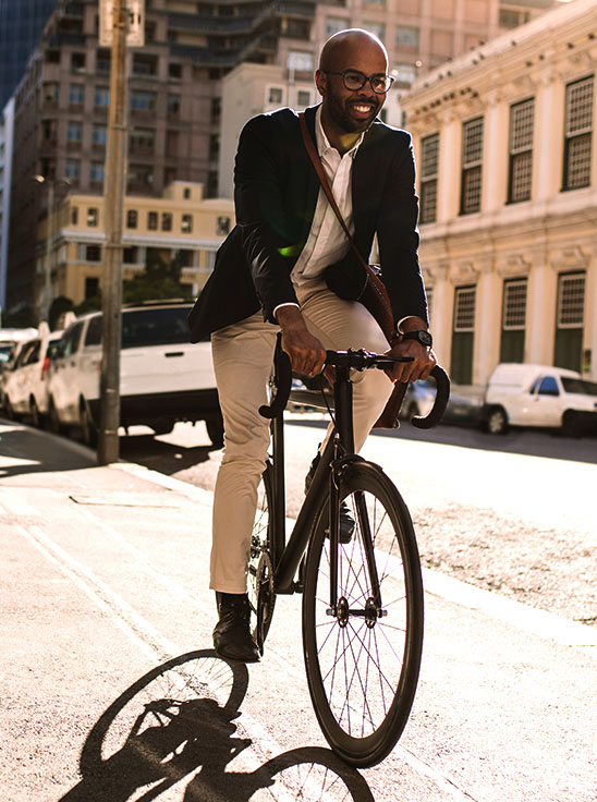A businessperson wearing a suit jacket, slacks, and dress shoes riding a bicycle on a city street sidewalk, and an aerial view of a busy city interstate and side streets