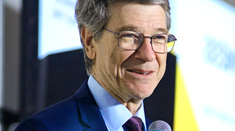 Economist Jeffery Sachs smiling, wearing thin square frame glasses, a blue shirt, and black blazer