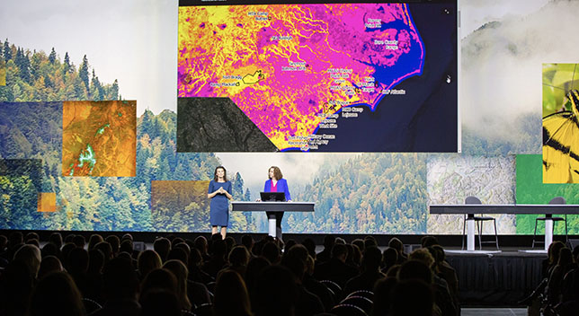 Two presenters on stage in a full room with a screen showing a bright pink and yellow map and background setting of tree