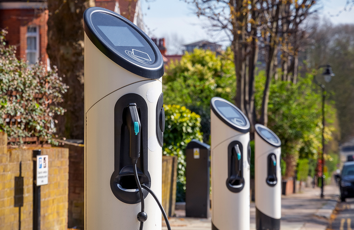 A row of electric vehicle charging stations along the sidewalk of a tree-lined street with brick buildings visible in the background