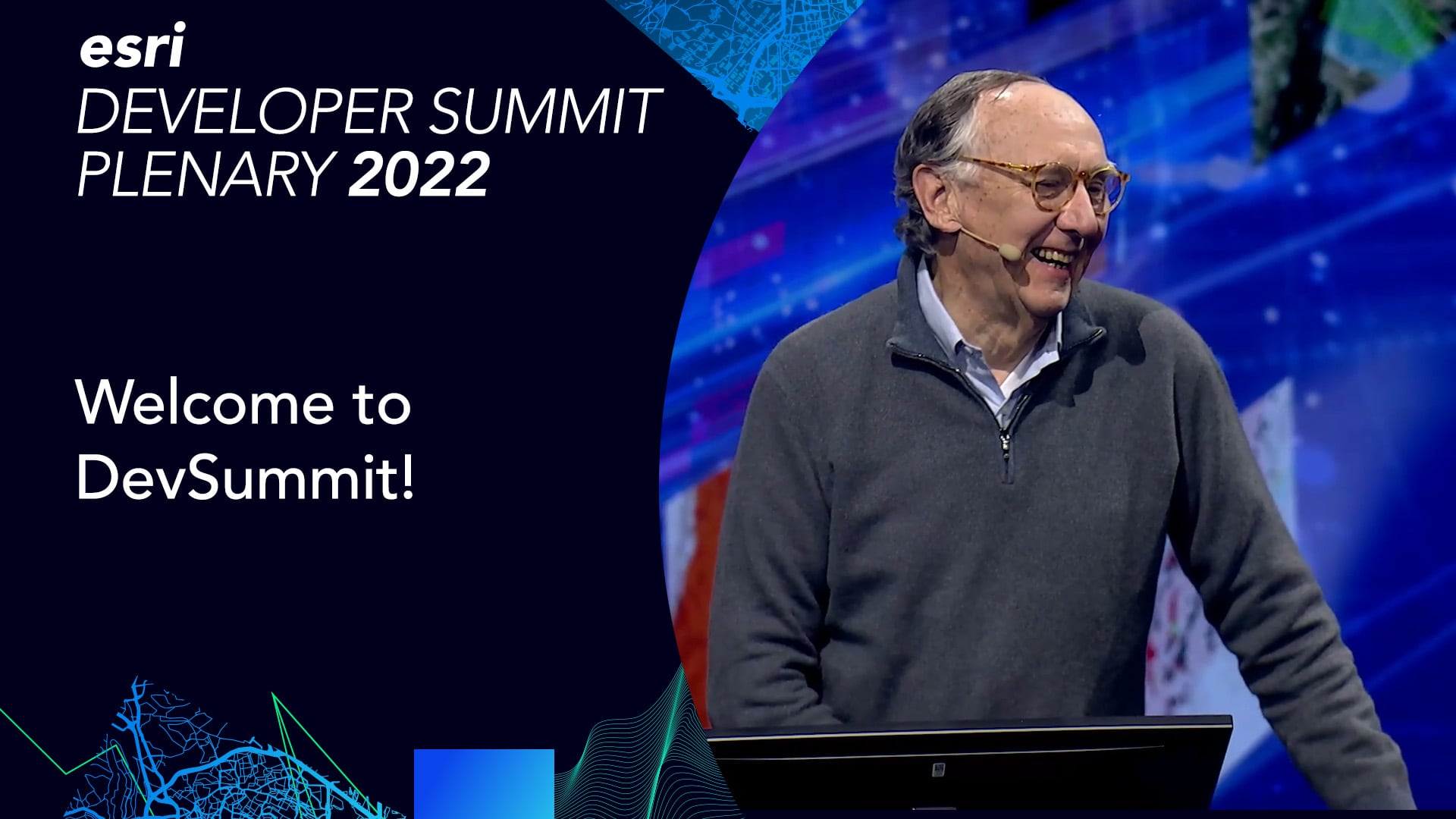 Jack Dangermond smiles while standing at a podium with the words, Esri Developer Summit Plenary 2022 and Welcome to DevSummit to the left