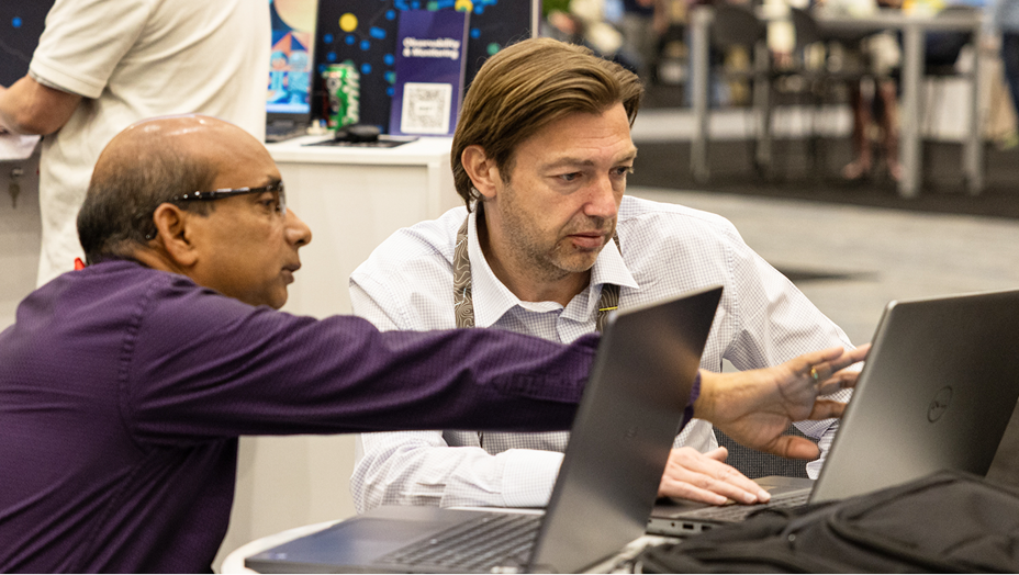 Two men sit side by side at a table, focusing on a laptop screen as one points and explains something to the other