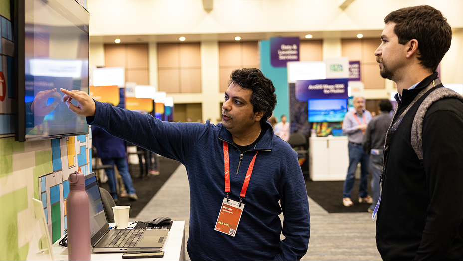 A man wearing a conference badge points at a large screen while speaking to another attendee