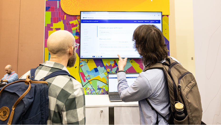 Two people stand in front of a large wall-mounted monitor at a conference or expo