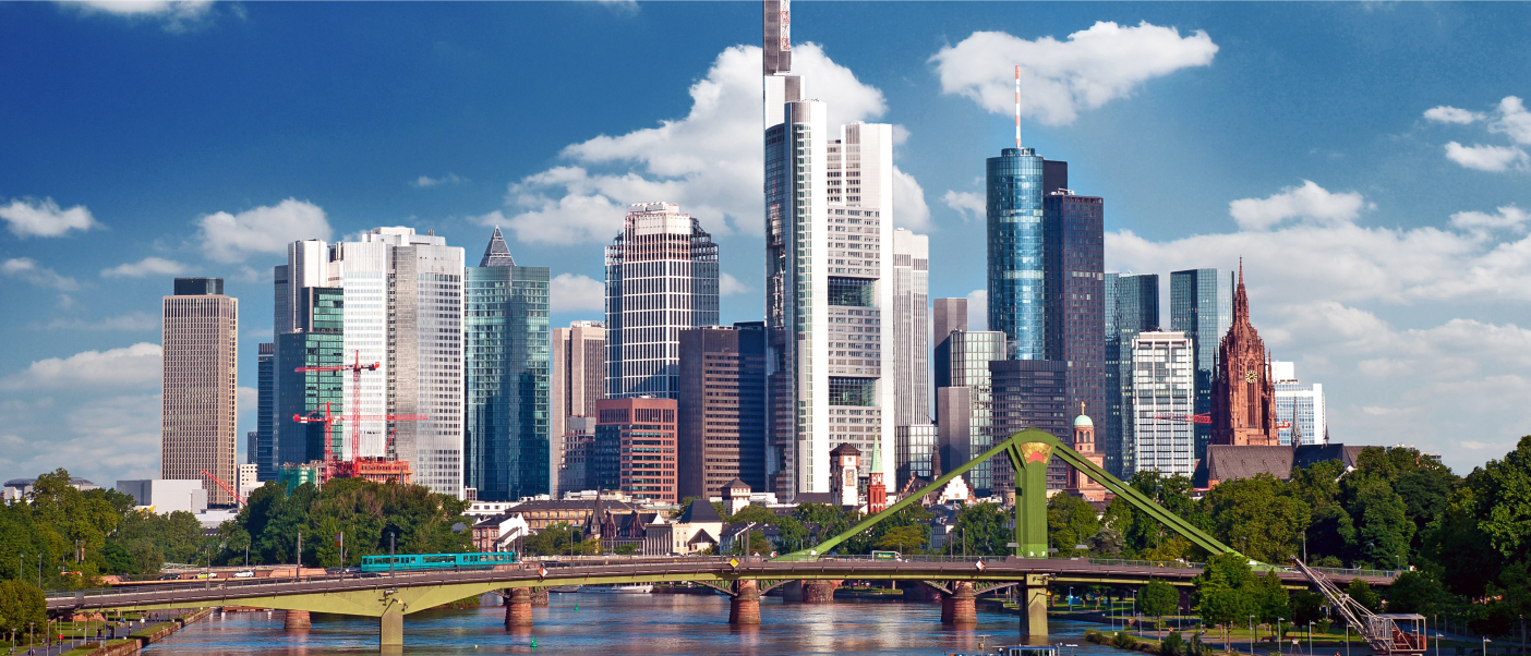 Frankfurt cityscape under a blue sky with scattered clouds and a river in the foreground crossed by a green bridge