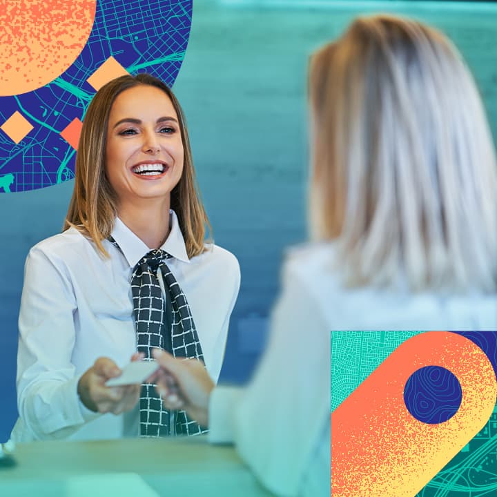 Smiling hotel employee dressed in a white blouse and black-patterned scarf hands a guest a hotel key at check in