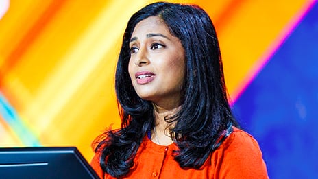 Presenter with long dark hair wearing an orange top, standing at a lectern against a vibrant background of yellow, orange, and blue diagonal lines