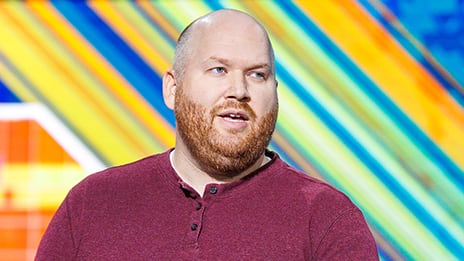 Presenter wearing a maroon long-sleeve henley shirt, positioned in front of a colorful background with diagonal yellow, blue, and green stripes