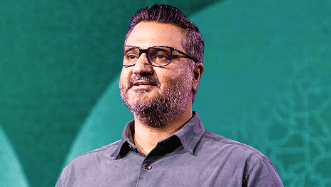 Portrait of a speaker wearing glasses and a gray button-up shirt against a teal background.