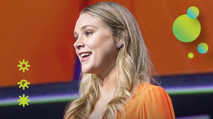 Presenter with long blonde curls, wearing an orange top, looks to the left while speaking