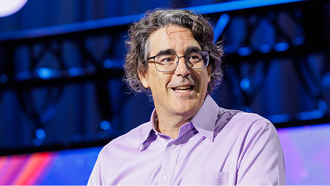 Presenter with gray hair, wearing a purple shirt, looks to the right while speaking onstage
