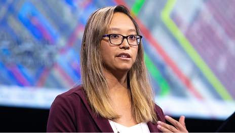Presenter with long hair and black-framed glasses gestures with one hand while speaking onstage