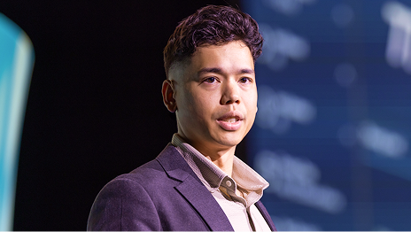 Presenter in a jacker and open-neck collared shirt onstage against a blurred background that is darker on the left