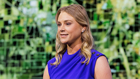 Blonde presenter wearing a bright blue top stands onstage against a green and black map
