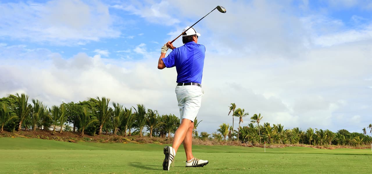 Golfer in shorts and a blue shirt on a golf course edged with palm trees, completing a backswing