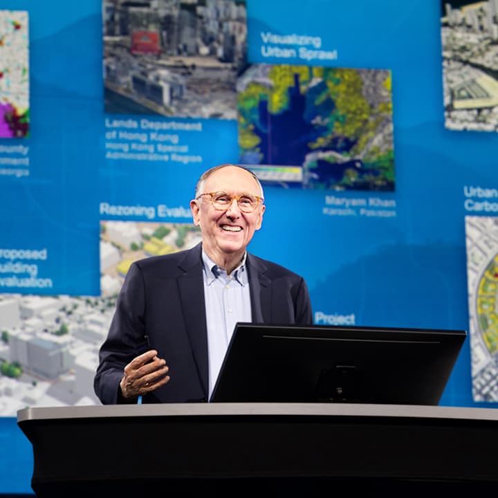 Jack Dangermond wearing a blue dress shirt and blazer on stage behind a podium with colorful maps and visualizations behind him