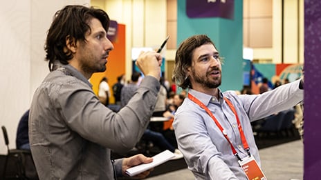 Two people standing at an expo booth with one pointing to a display screen while explaining content