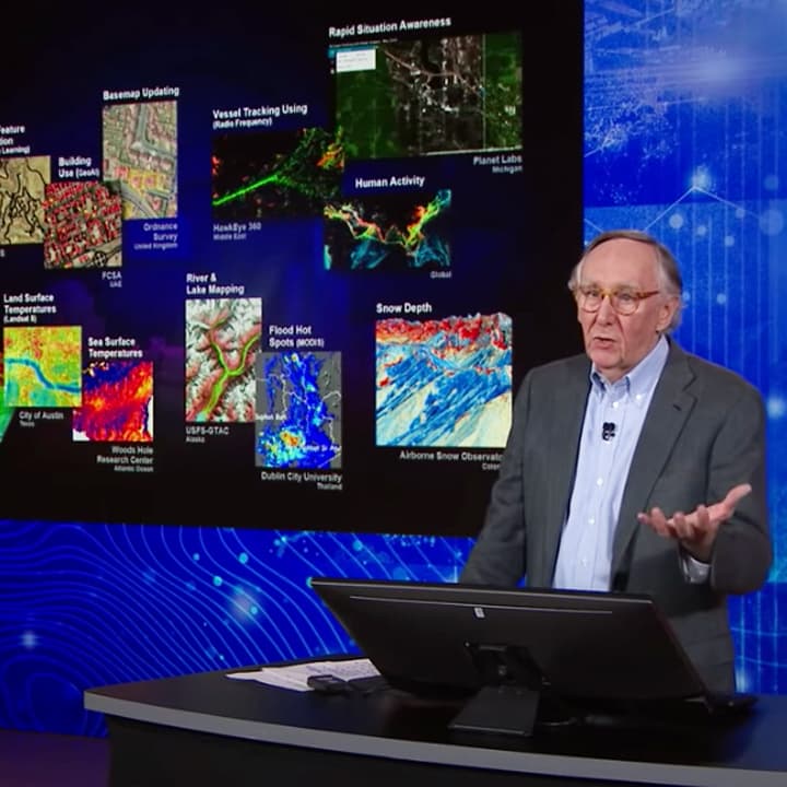 Jack Dangermond speaking at a podium with a variety of colorful user maps and images displayed behind him on the left