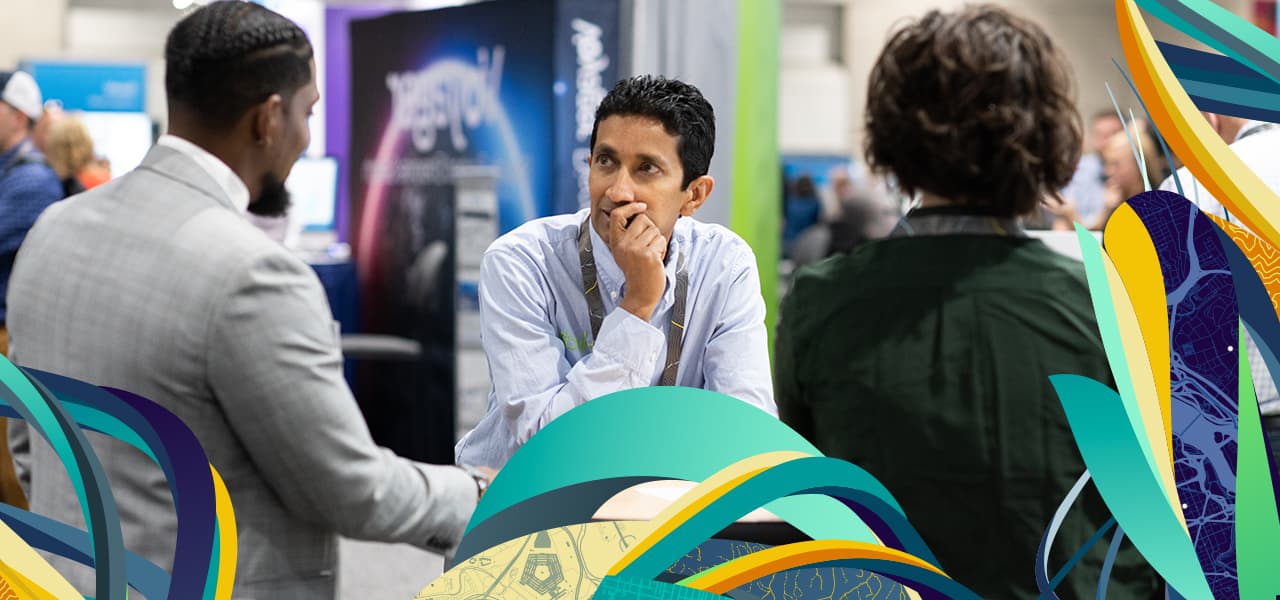 A trade show attendee in an exhibit booth looks thoughtful as another person speaks; a third person stands on the right