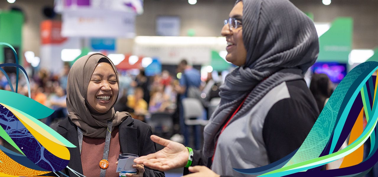 Two women in headscarves at a trade show; one gestures with her hands while the other is laughing, eyes closed