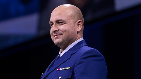 Bald speaker in a dark blue military dress uniform onstage during a presentation