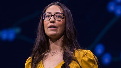 Presenter with long dark hair and glasses looks slightly to the left while speaking onstage.