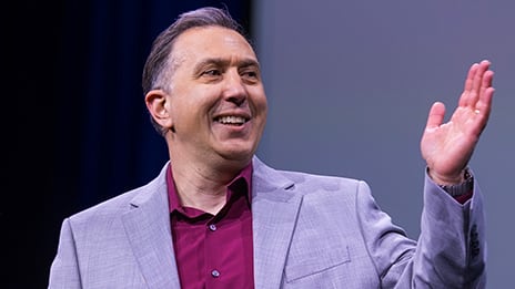 Speaker in a light purple jacket smiles and gestures with one hand during a presentation