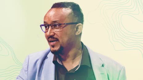 A man wearing a light blue blazer and dark shirt is captured against a pastel abstract background featuring contour-like patterns. 