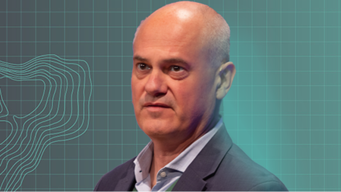 Portrait of a speaker wearing a grey suit jacket against a blue-green gradient background with a grid and contour map lines