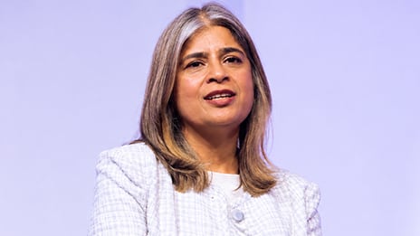 A middle-aged woman is shown speaking, captured from the chest up against a soft purple background