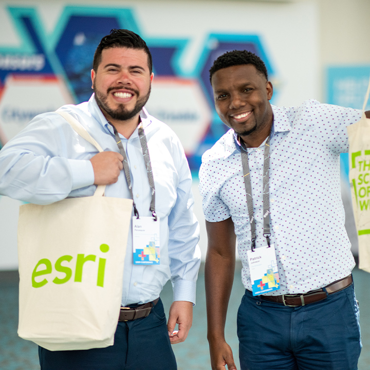 Two conference attendees smiling at the camera while they wear their badges and one wears an Esri swag bag over his shoulder