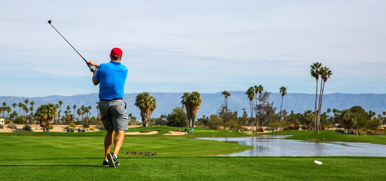 Rear view of a golfer just after a shot, on a course with water, palm trees, and mountains in the background